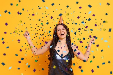 Image of Happy woman in conical paper hat under falling confetti on orange background. Surprise party