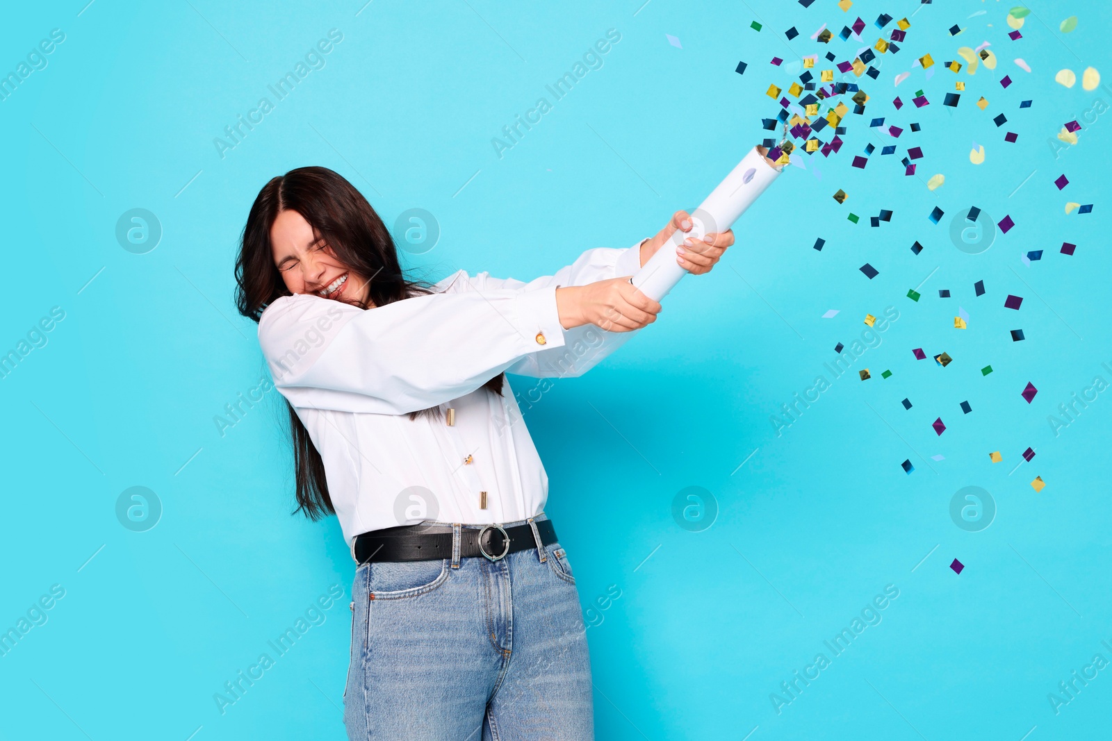 Image of Happy woman blowing up confetti popper on light blue background. Surprise party