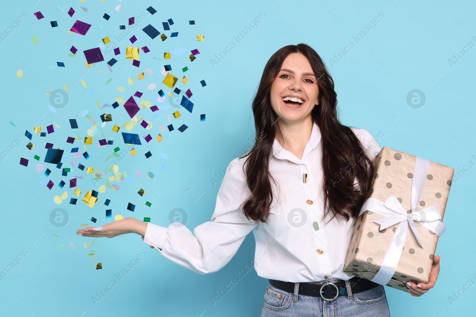 Image of Happy woman with gift and falling confetti on light blue background. Surprise party