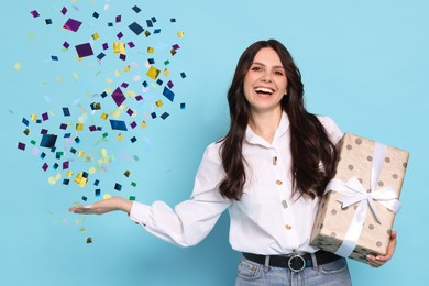 Happy woman with gift and falling confetti on light blue background. Surprise party