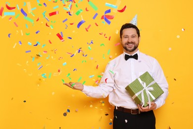 Image of Happy man in conical paper hat with gift under falling confetti on orange background. Surprise party