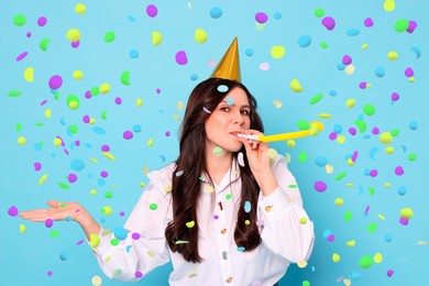 Image of Beautiful woman in conical paper hat with blower under falling confetti on light blue background. Surprise party