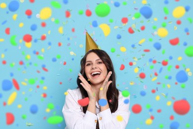 Image of Happy woman in conical paper hat under falling confetti on light blue background. Surprise party