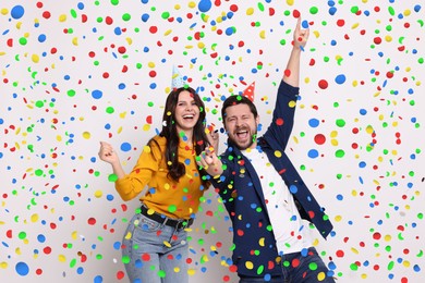 Image of Happy woman and man in conical paper hats under falling confetti on light background. Surprise party