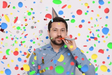 Image of Man in conical paper hat with blower under falling confetti on light background. Surprise party