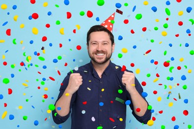 Happy man in conical paper hat under falling confetti on light blue background. Surprise party