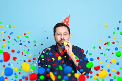 Image of Man in conical paper hat with blower and falling confetti on light blue background. Surprise party