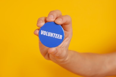 Man holding button badge with word Volunteer on orange background, closeup