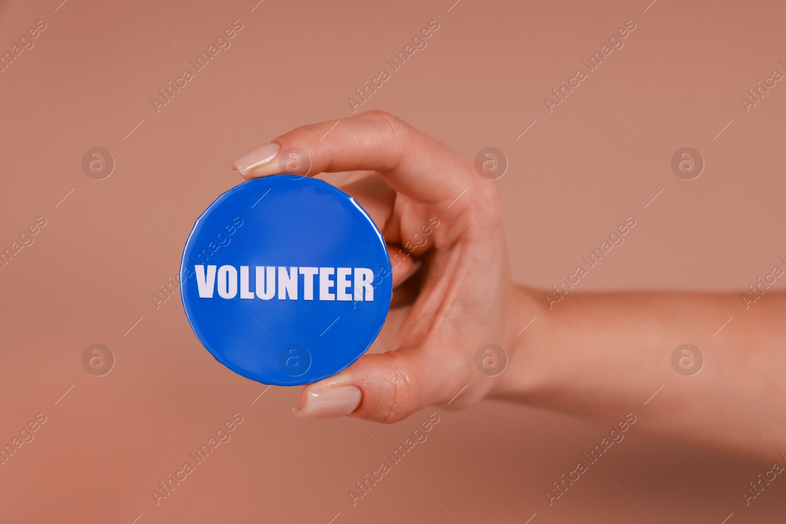 Photo of Woman showing blue button badge with word Volunteer on light brown background, closeup