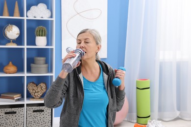 Photo of Elderly woman drinking water during exercise with dumbbell at home