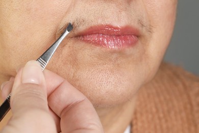 Photo of Senior woman plucking her mustache with tweezers on grey background, closeup