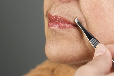 Photo of Senior woman plucking her mustache with tweezers on grey background, closeup