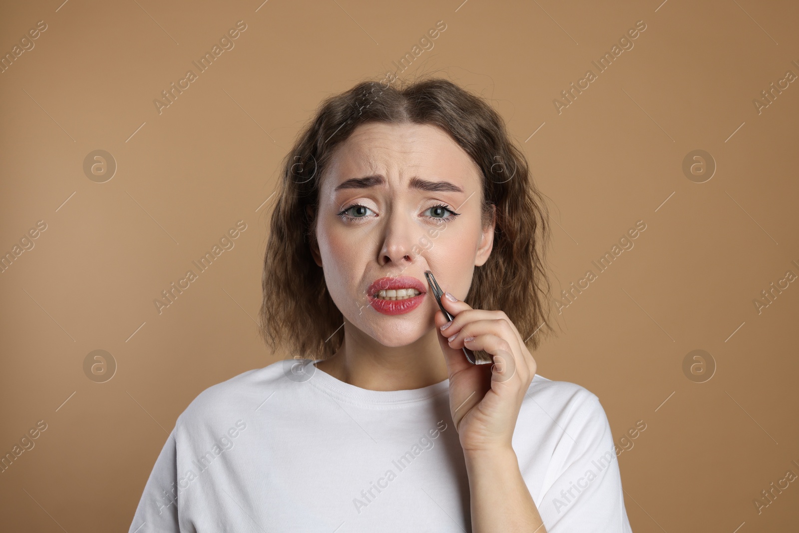 Photo of Disappointed woman plucking her mustache with tweezers on beige background