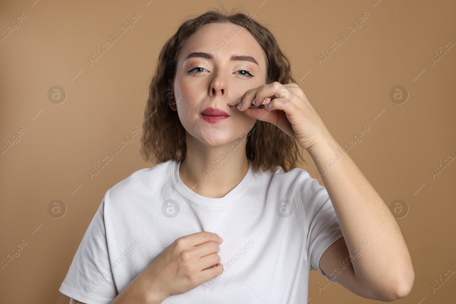 Photo of Beautiful woman plucking her mustache with tweezers on beige background