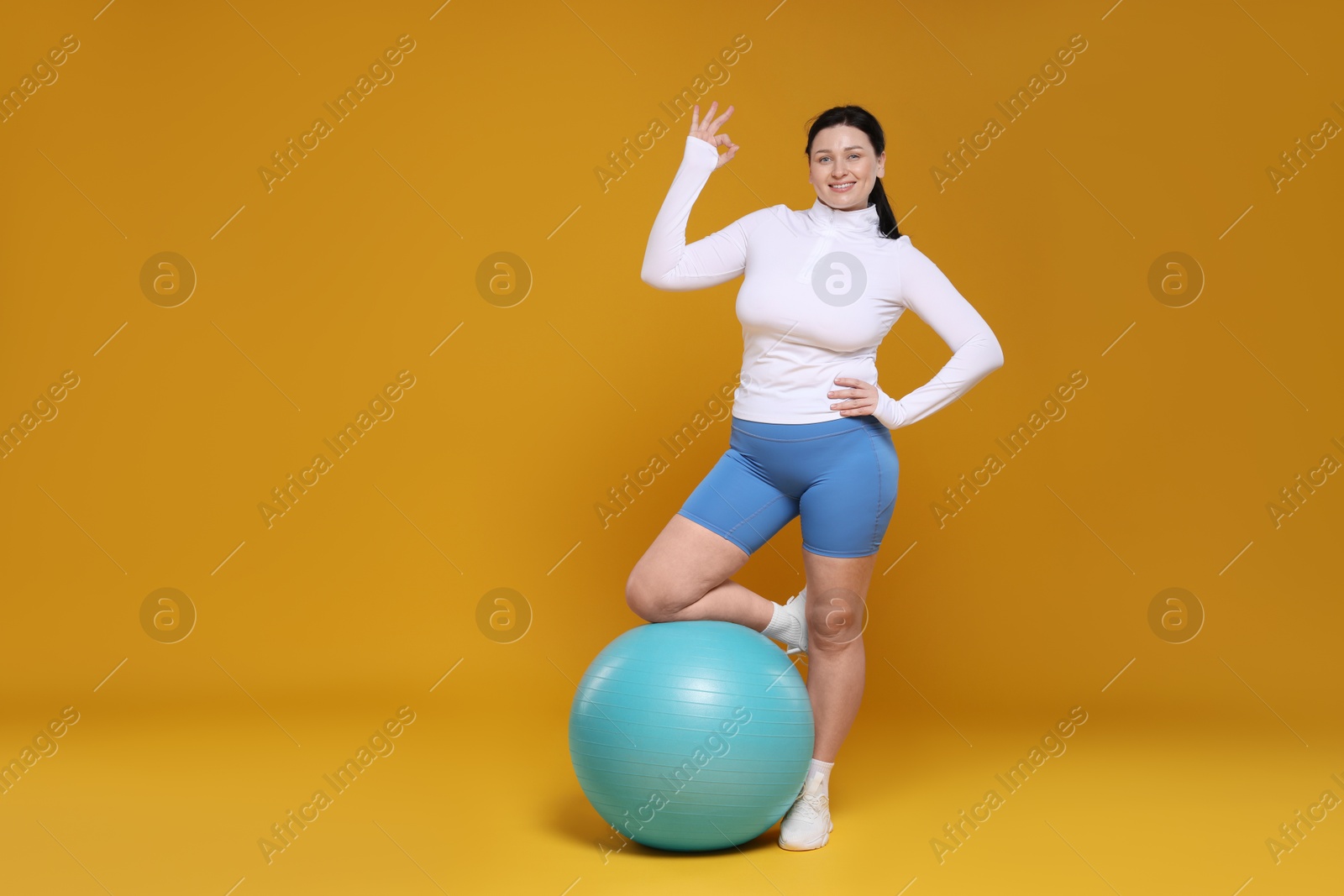 Photo of Plus size woman in gym clothes with fitness ball showing ok gesture on orange background, space for text