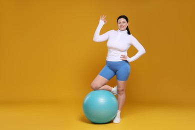 Photo of Plus size woman in gym clothes with fitness ball showing ok gesture on orange background, space for text