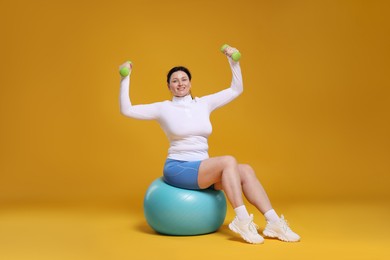 Plus size woman in gym clothes with dumbbells and fitness ball on orange background