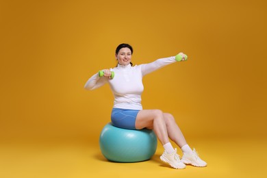 Plus size woman in gym clothes with dumbbells and fitness ball on orange background