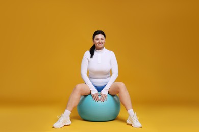 Plus size woman in gym clothes with fitness ball on orange background