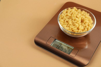 Photo of Electronic kitchen scale with bowl of raw pasta on beige background, closeup. Space for text