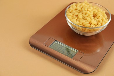 Photo of Electronic kitchen scale with bowl of raw pasta on beige background, closeup. Space for text