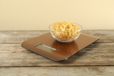 Photo of Electronic kitchen scale with bowl of raw pasta on wooden table against beige background, closeup