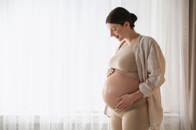 Beautiful pregnant woman near window at home. Space for text