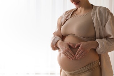 Pregnant woman making heart with hands on her belly near window at home, closeup. Space for text