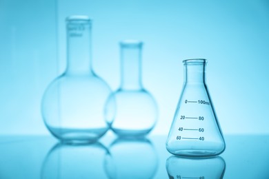 Photo of Empty flasks on mirror surface against light blue background. Laboratory equipment