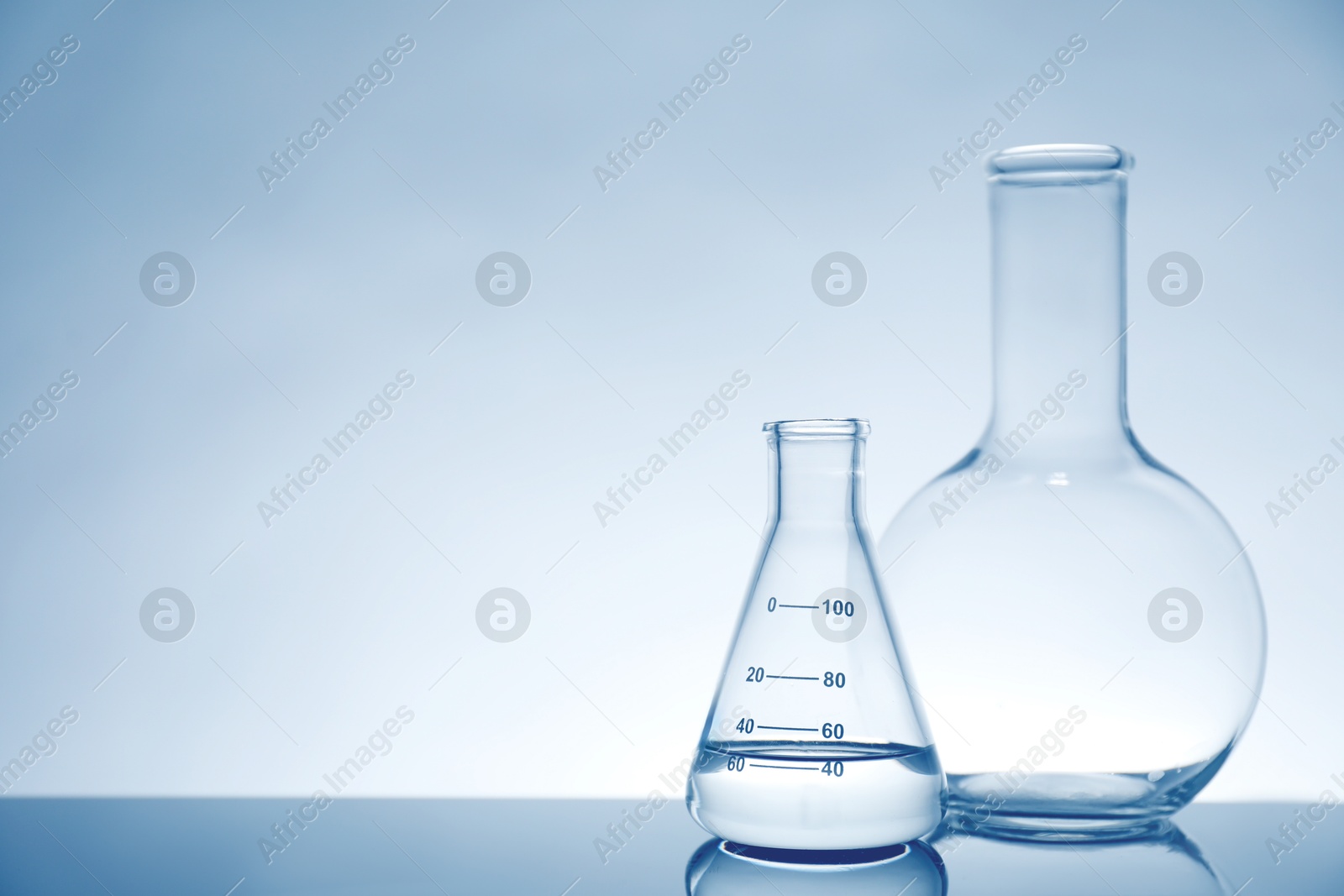 Photo of Laboratory glassware with liquid on mirror surface against light background, space for text