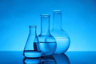 Photo of Flasks with liquid on mirror surface against light blue background. Laboratory equipment