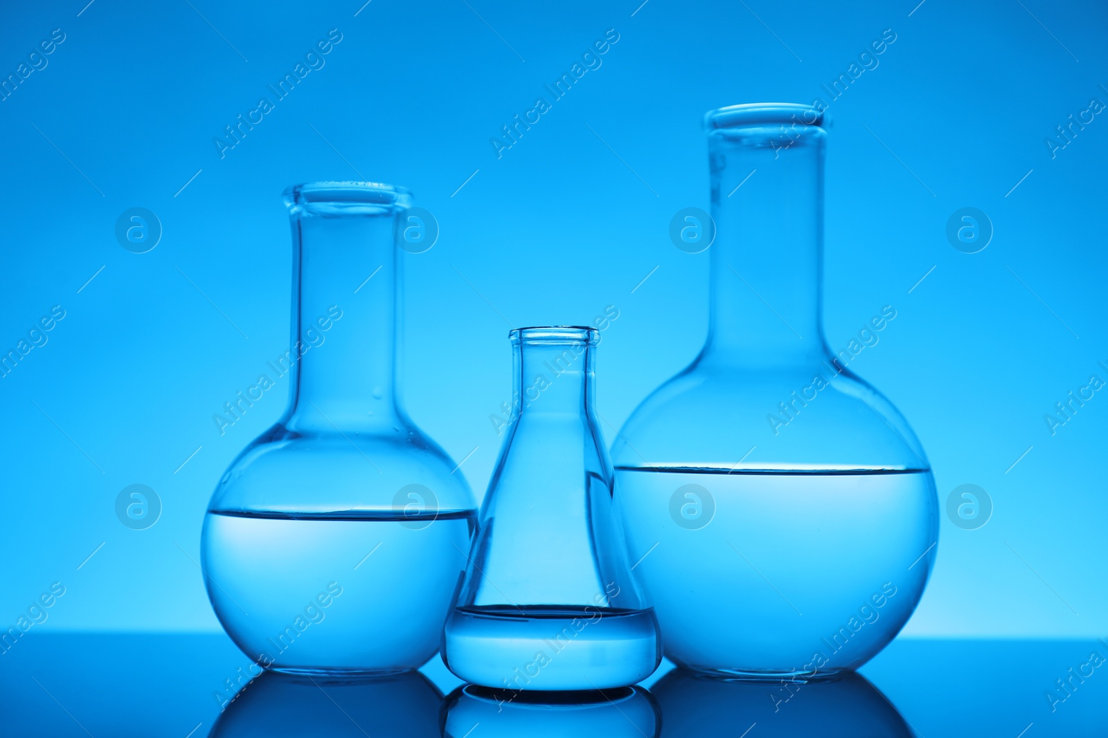 Photo of Flasks with liquid on mirror surface against light blue background. Laboratory equipment