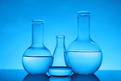 Flasks with liquid on mirror surface against light blue background. Laboratory equipment