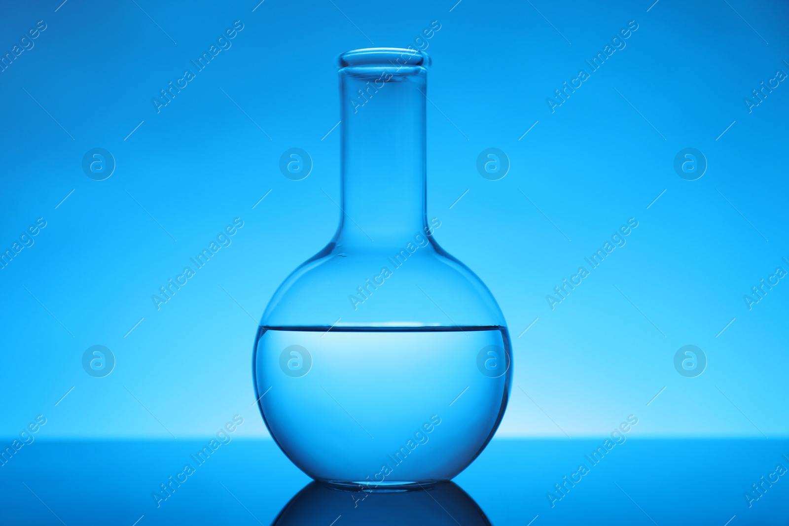 Photo of Flask with liquid on mirror surface against light blue background. Laboratory equipment