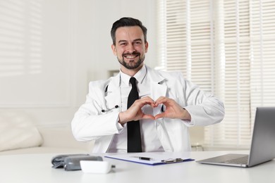 Photo of Smiling cardiologist making heart with hands at white table in clinic