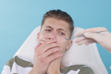 Scared man covering mouth and dentist with tool on light blue background