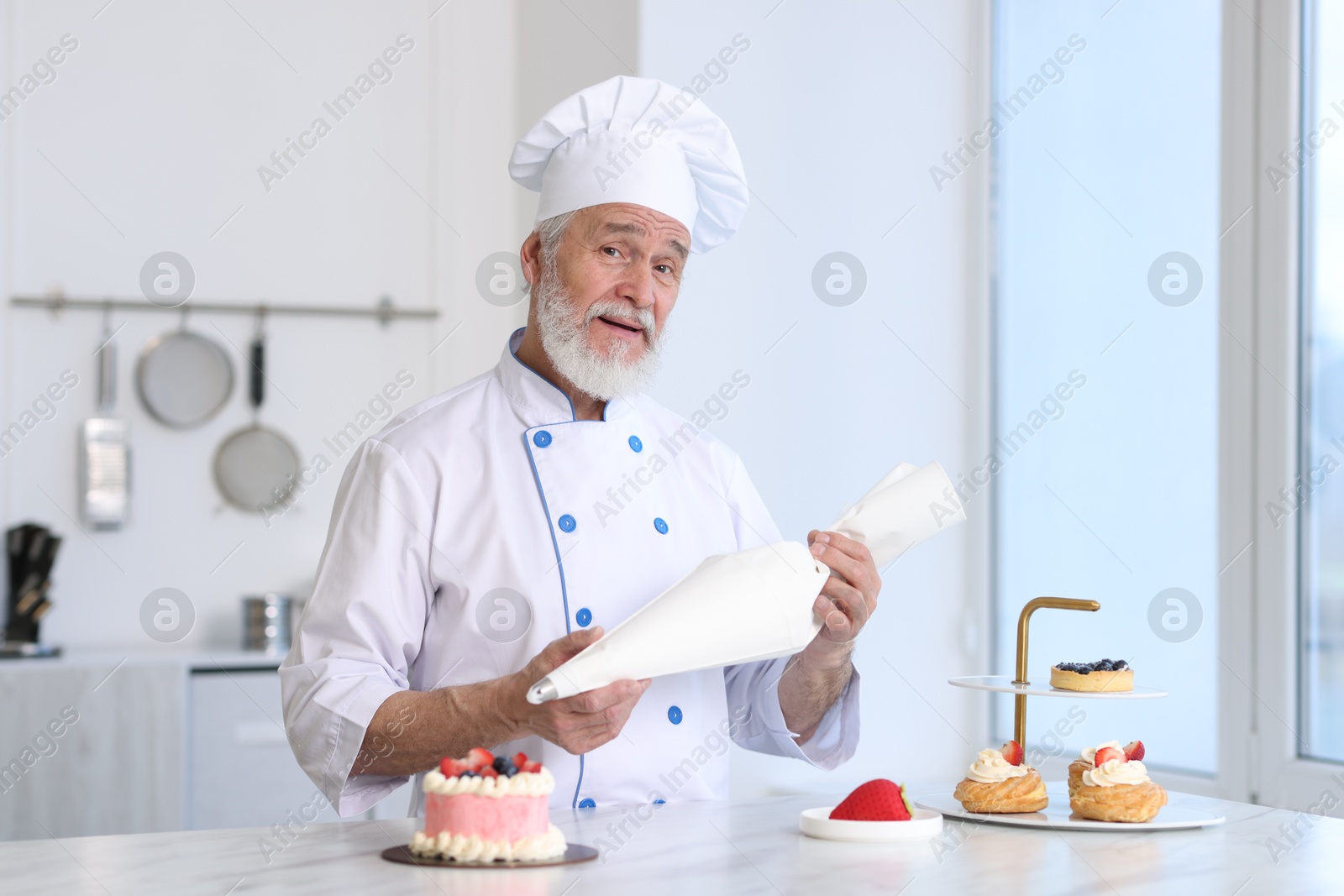 Photo of Elderly confectioner with pastry bag making cake at table in kitchen