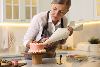 Photo of Senior confectioner with pastry bag making cake at table in kitchen