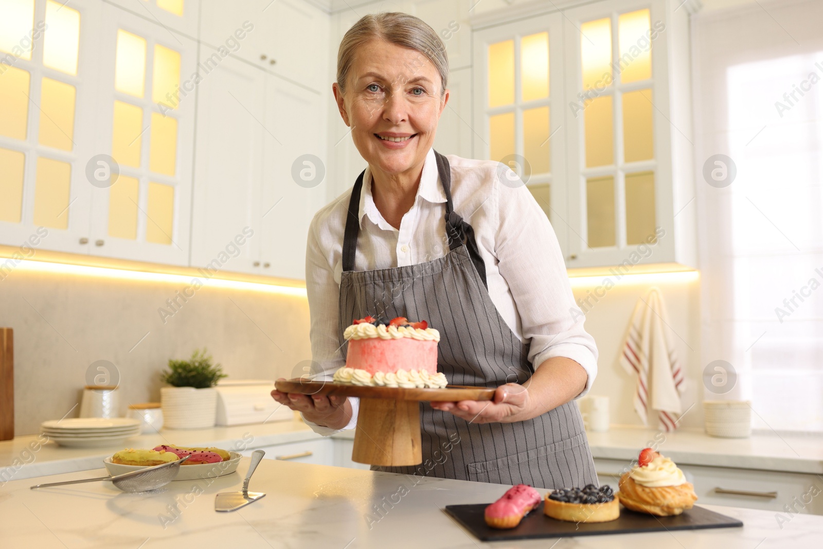 Photo of Smiling pastry chef holding tasty cake at table with desserts in kitchen