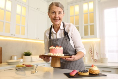 Smiling pastry chef holding tasty cake at table with desserts in kitchen