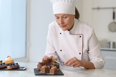 Photo of Senior pastry chef decorating tasty desserts with edible gold leaf sheet at table in kitchen