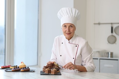 Photo of Smiling pastry chef decorating tasty desserts with edible gold leaf sheet at table in kitchen