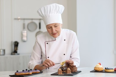 Senior pastry chef decorating tasty desserts with edible gold leaf sheet at table in kitchen