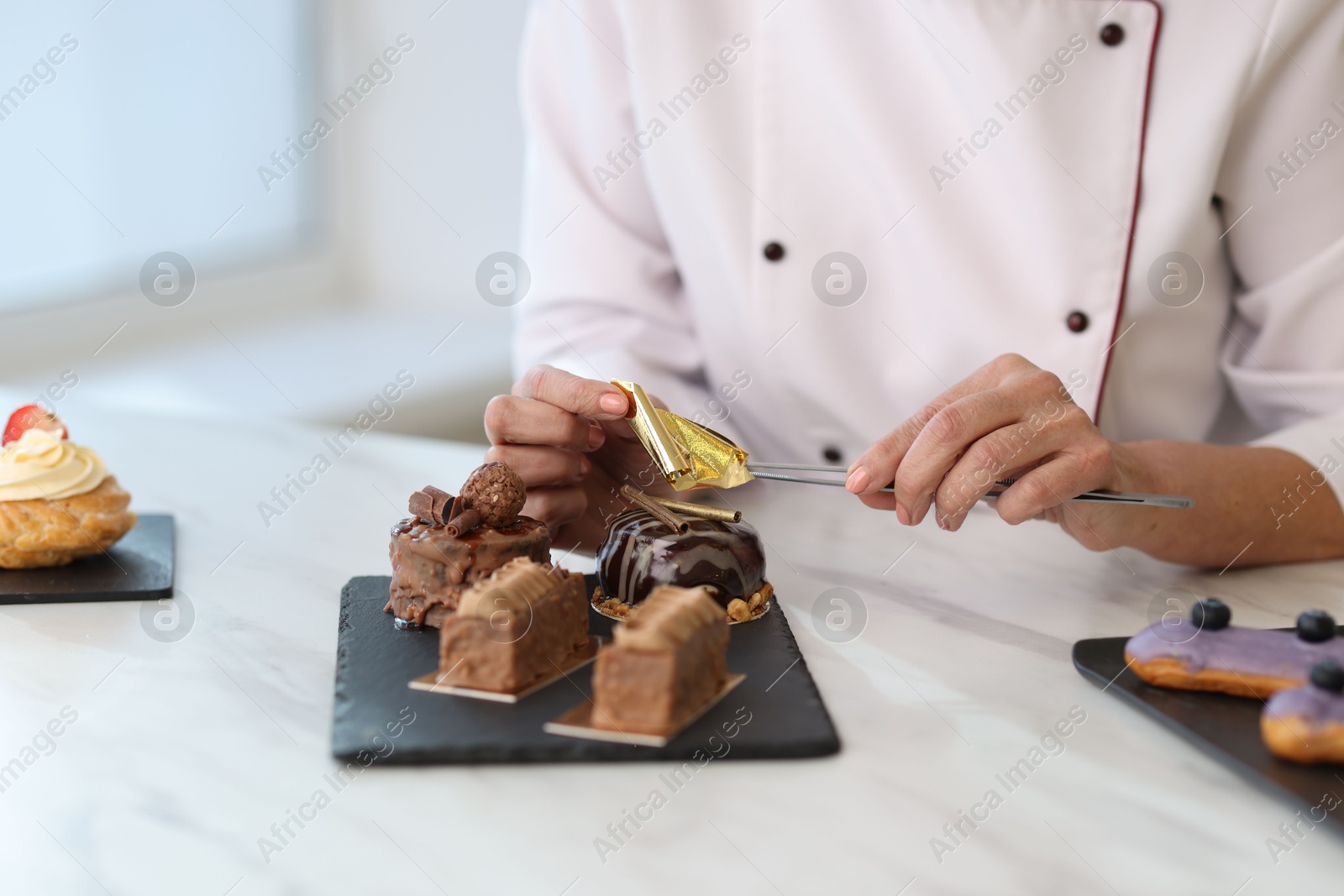 Photo of Pastry chef decorating tasty desserts with edible gold leaf sheet at table in kitchen, closeup