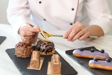 Photo of Pastry chef decorating tasty desserts with edible gold leaf sheet at table in kitchen, closeup