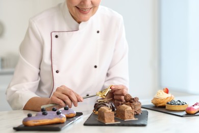 Smiling pastry chef decorating tasty desserts with edible gold leaf sheet at table in kitchen, closeup