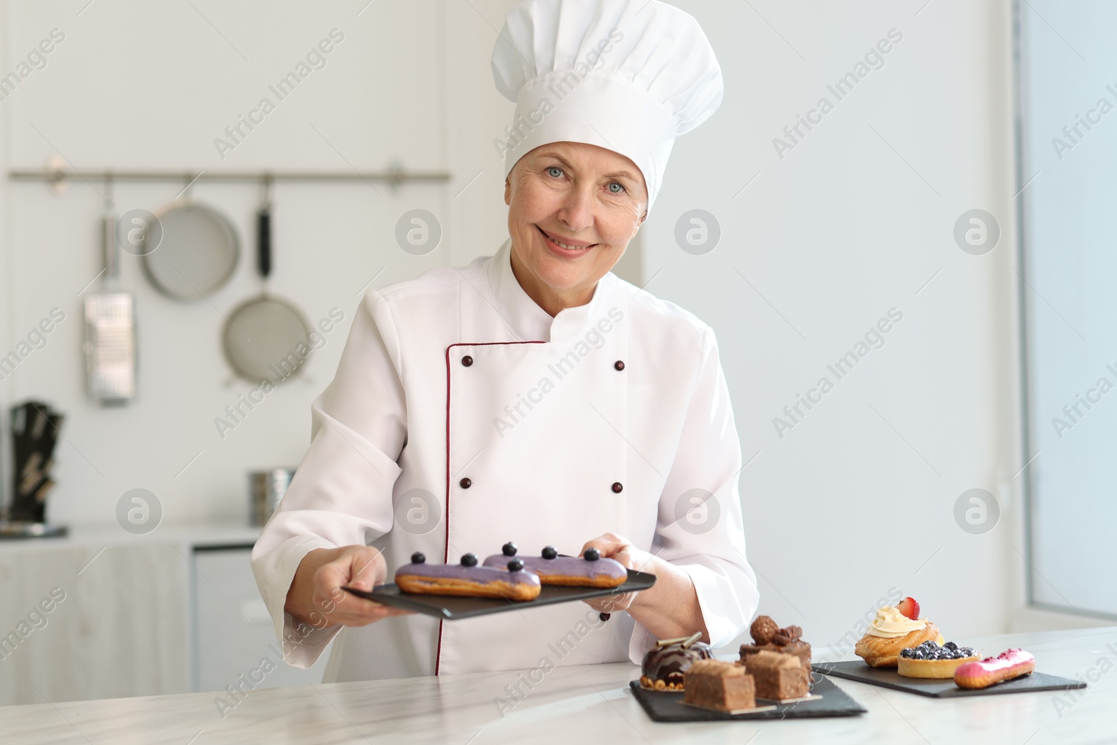 Photo of Smiling pastry chef holding tasty eclairs at table with other desserts in kitchen