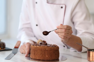 Photo of Pastry chef decorating cake with melted chocolate at table in kitchen, closeup