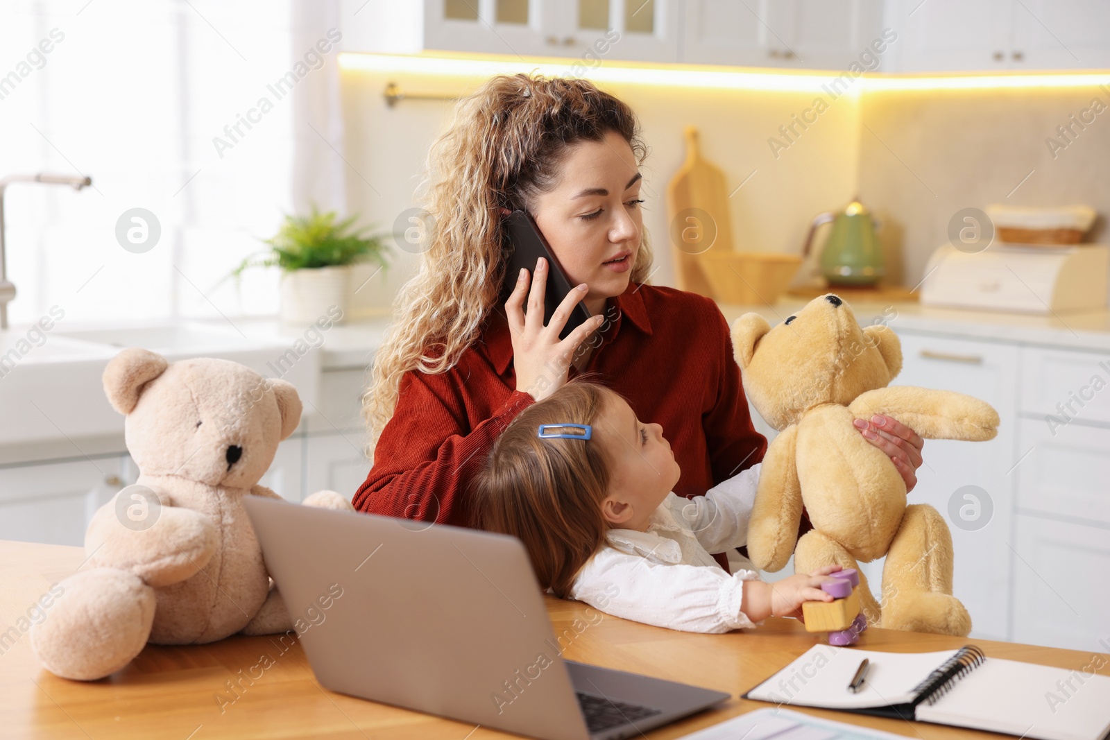 Photo of Work-family balance. Single mother holding her daughter while talking on smartphone at table in kitchen