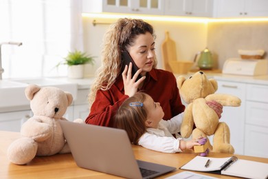 Photo of Work-family balance. Single mother holding her daughter while talking on smartphone at table in kitchen